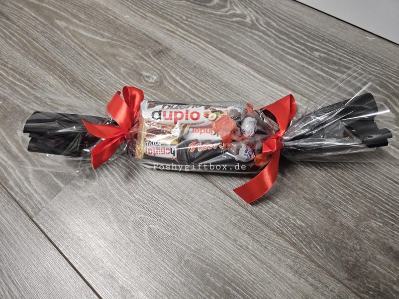 May include: A clear cellophane gift wrapper shaped like a large sweet, containing assorted chocolate bars including Duplo and Kinder products. The ends are twisted and secured with bright red satin ribbons. The text poshygiftbox.de is visible.