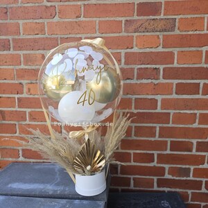 May include: A clear balloon filled with white and gold balloons and white confetti, featuring the gold text Rahmiye and 40. The balloon is attached to a white cylindrical base decorated with dried pampas grass and a gold fan-shaped ornament.