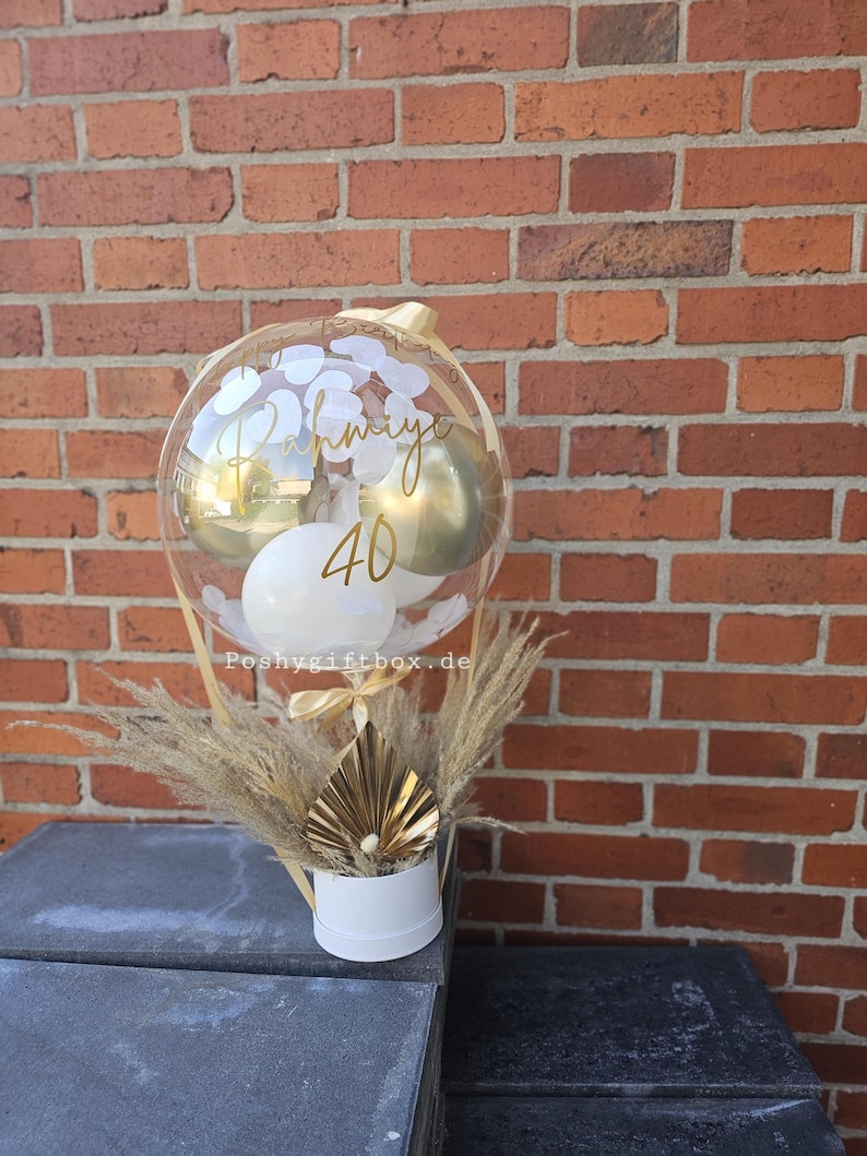 May include: A clear balloon filled with white and gold balloons and white confetti, featuring the gold text Rahmiye and 40. The balloon is attached to a white cylindrical base decorated with dried pampas grass and a gold fan-shaped ornament.