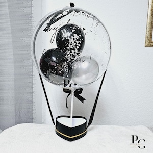 May include: A clear balloon hot air balloon with black and silver balloons inside. The balloon is tied to a black heart-shaped box with a gold trim. The balloon has the text "Happy 33rd Birthday my love you!" written on it.
