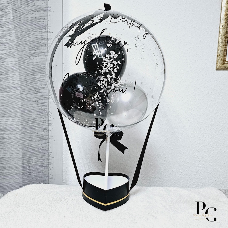 May include: A clear balloon hot air balloon with black and silver balloons inside. The balloon is tied to a black heart-shaped box with a gold trim. The balloon has the text "Happy 33rd Birthday my love you!" written on it.