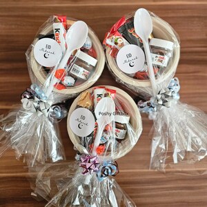 May include: Three clear plastic bags filled with various chocolate candies, a small jar of Nutella, a white plastic spoon, and a white circle sticker with the text "Eid Mubarak" and a crescent moon. The bags are tied with a ribbon and a bow.