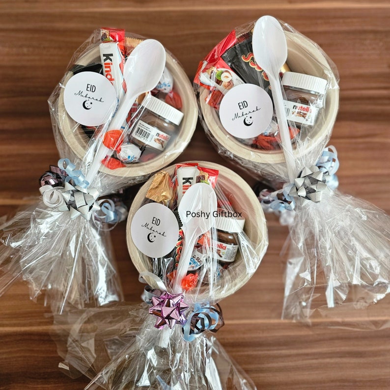 May include: Three clear plastic bags filled with various chocolate candies, a small jar of Nutella, a white plastic spoon, and a white circle sticker with the text "Eid Mubarak" and a crescent moon. The bags are tied with a ribbon and a bow.