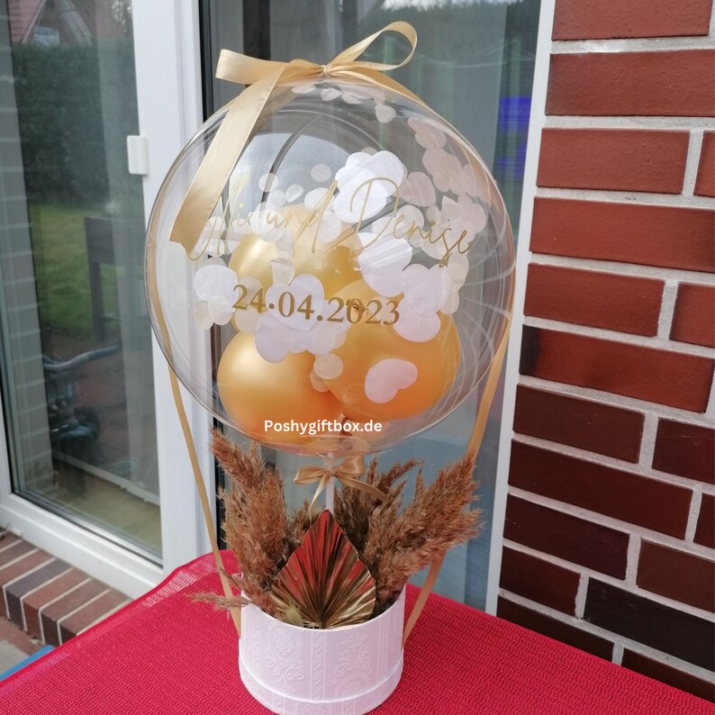 May include: A clear balloon filled with smaller gold balloons and white confetti, featuring the gold text "Miriam Denise" and "24.04.2023". The balloon is attached to a white cylindrical box containing dried pampas grass and a gold fan.