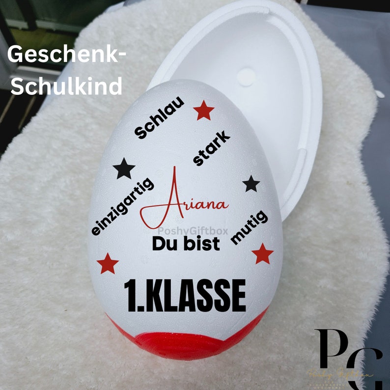 Peut inclure: Conteneur blanc en forme d'&oelig;uf avec un fond rouge et du texte noir qui dit "Geschenk-Schulkind", "Schlau", "stark", "einzigartig", "Ariana", "Du bist", "mutig", "1.KLASSE". Il y a des &eacute;toiles rouges et noires autour du texte.