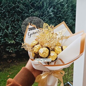 May include: A white and gold gift box bouquet with a clear acrylic sign that says "F&uuml;r Erna zum 80. Geburtstag". The bouquet is filled with chocolate truffles and dried flowers.