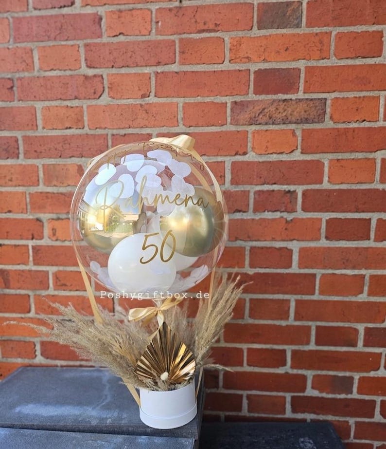 May include: A clear balloon filled with smaller white and gold balloons, featuring the gold text "Rahmena" and "50" on the surface. The balloon is attached to a white cylindrical base decorated with dried pampas grass and a gold fan-shaped accent.
