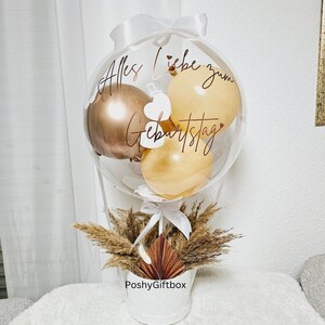 May include: A clear balloon filled with smaller metallic gold and beige balloons, featuring the text Alles Liebe zum Geburtstag in dark script. The balloon is tied with a white ribbon and sits in a white container decorated with dried pampas grass.