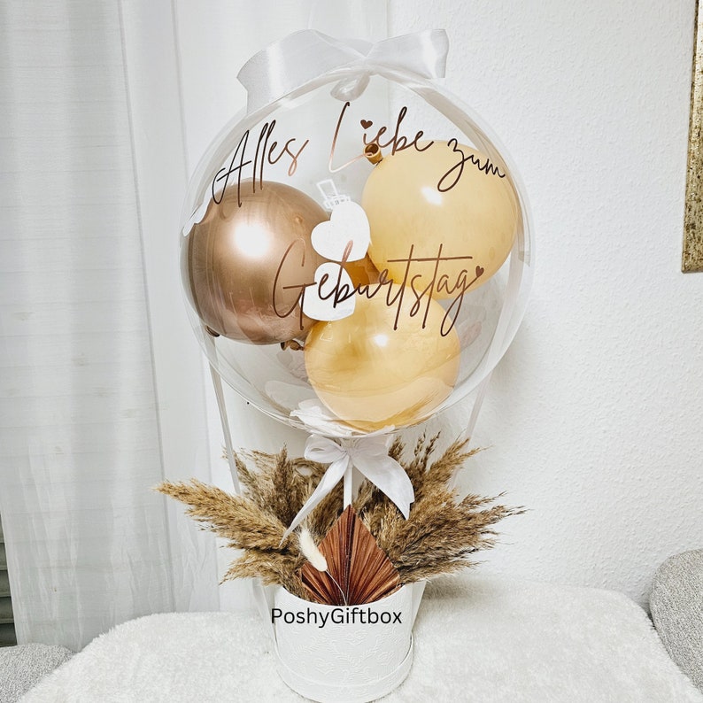 May include: A clear balloon filled with smaller metallic gold and beige balloons, featuring the text Alles Liebe zum Geburtstag in dark script. The balloon is tied with a white ribbon and sits in a white container decorated with dried pampas grass.