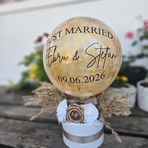 以下が含まれることがあります： 金色のマーブル模様が施された球体のバルーン。黒い文字で「JUST MARRIED」「Ebru & Stefan」「09.06.2026」と印字されています。白い円筒形のボックスに収められ、白と金の布製のバラと乾燥したパンパスグラスで装飾されています。