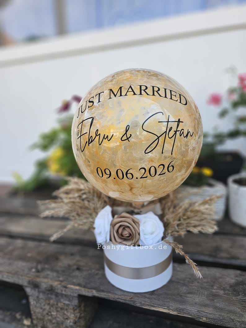 以下が含まれることがあります： 金色のマーブル模様が施された球体のバルーン。黒い文字で「JUST MARRIED」「Ebru & Stefan」「09.06.2026」と印字されています。白い円筒形のボックスに収められ、白と金の布製のバラと乾燥したパンパスグラスで装飾されています。
