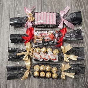 May include: Four candy gift crackers wrapped in black cellophane with clear plastic, tied with pink, red, and gold ribbons. Each contains assorted chocolates and sweets, including pink wafer sticks, Duplo, Ferrero Rocher, and Raffaello candies.