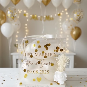 May include: A clear box filled with white balloons and gold confetti, with the words "JUST MARRIED" and names and a date in gold lettering. The box is decorated with a white ribbon, a white rose, and dried flowers. Gold and white balloons are in the background.