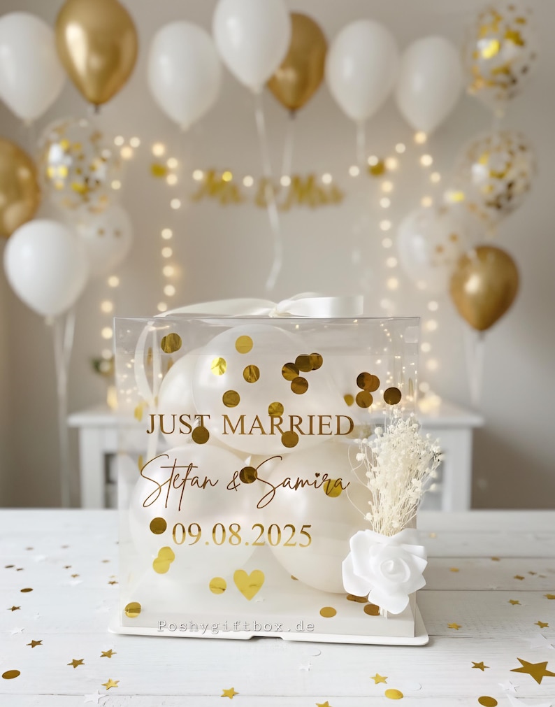 May include: A clear box filled with white balloons and gold confetti, with the words "JUST MARRIED" and names and a date in gold lettering. The box is decorated with a white ribbon, a white rose, and dried flowers. Gold and white balloons are in the background.