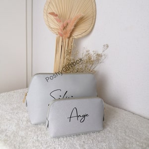 May include: Two gray zippered cosmetic bags with gold zippers. The larger bag has the name "Ellie" written in black cursive. The smaller bag has the name "Angie" written in black cursive.