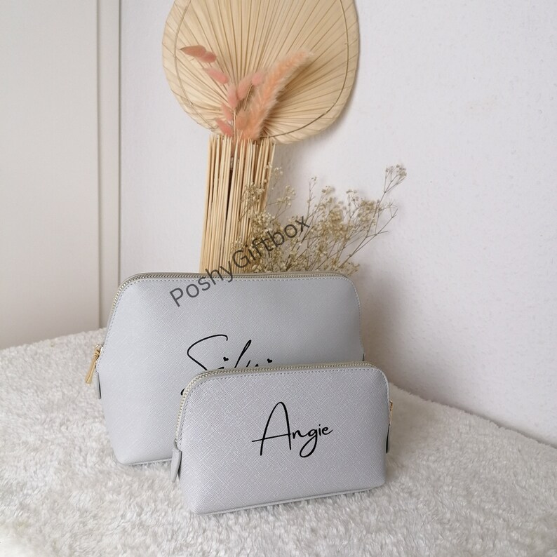 May include: Two gray zippered cosmetic bags with gold zippers. The larger bag has the name "Ellie" written in black cursive. The smaller bag has the name "Angie" written in black cursive.