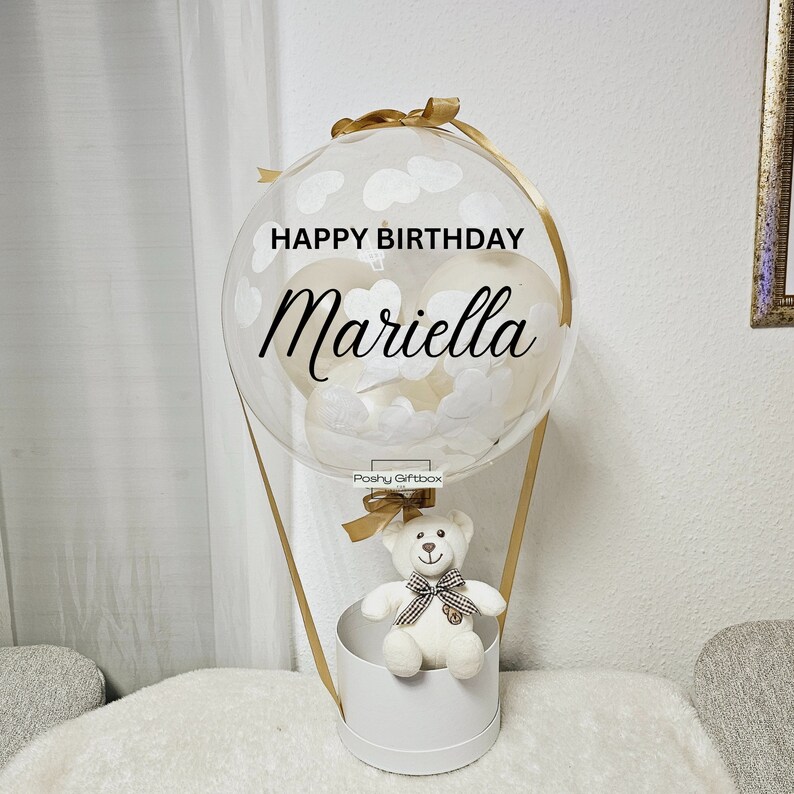 May include: A white teddy bear sits in a white box with a gold ribbon. The box is attached to a clear balloon with white hearts and clouds. The balloon has the text "Happy Birthday Mariella" on it.