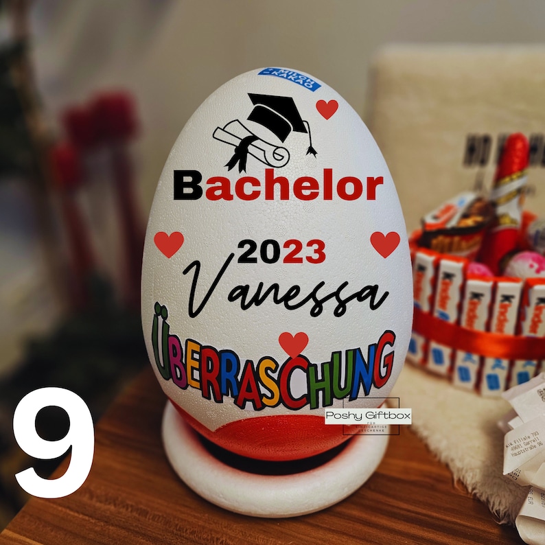 Może przedstawiać: Biały pojemnik w kształcie jajka z czapką doktorską, dyplomem i tekstem "Bachelor 2023 Vanessa &Uuml;berraschung" w kolorowych literach. Pojemnik jest ozdobiony czerwonymi sercami.