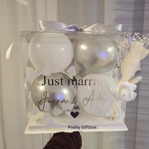 May include: A clear box with four balloons, two white and two silver, for a wedding. The box is decorated with a silver ribbon, dried flowers, and the words "Just married" and names. The box is labeled "Poshy Giftbox".