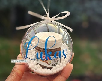 May include: A clear, spherical, hollow plastic bauble containing a miniature jar of Nutella surrounded by small white foam beads. The exterior of the bauble features the name Lukas in blue script lettering and a white ribbon bow at the top.