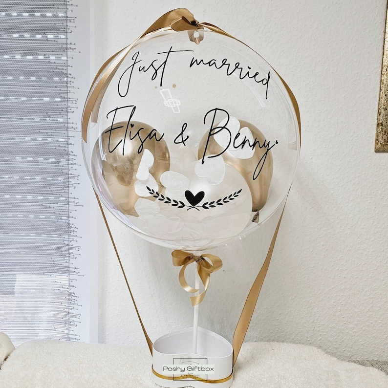 May include: A clear balloon hot air balloon with gold balloons inside. The balloon has a white ribbon and a white box with the text "Poshy Giftbox" on it. The balloon has the text "Just married Elisa & Benny" written on it.