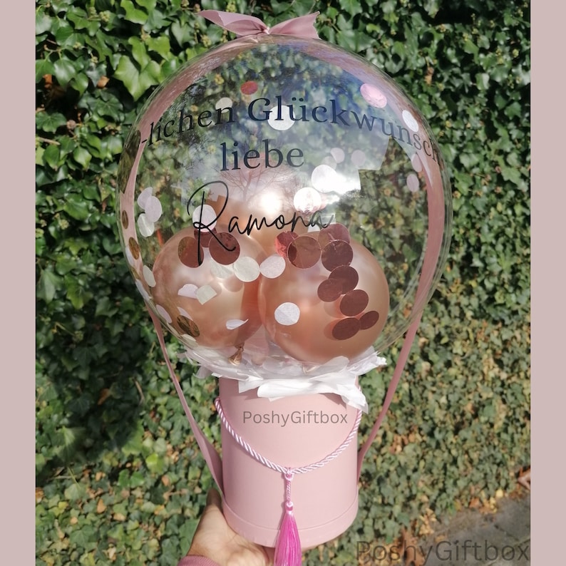 Op de afbeelding: Een doorzichtige ballon gevuld met confetti en drie gouden ballonnen, met de tekst "lichen Gl&uuml;ckwunsch liebe Ramona" op de ballon gedrukt. De ballon staat op een roze geschenkdoos met een kwast.
