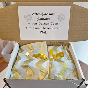May include: Open cardboard gift box with a white card that reads "Alles Gute zum Jubil&auml;um - von Deinem Team f&uuml;r einen besonderen Chef." The box is filled with gold confetti and white tissue paper.