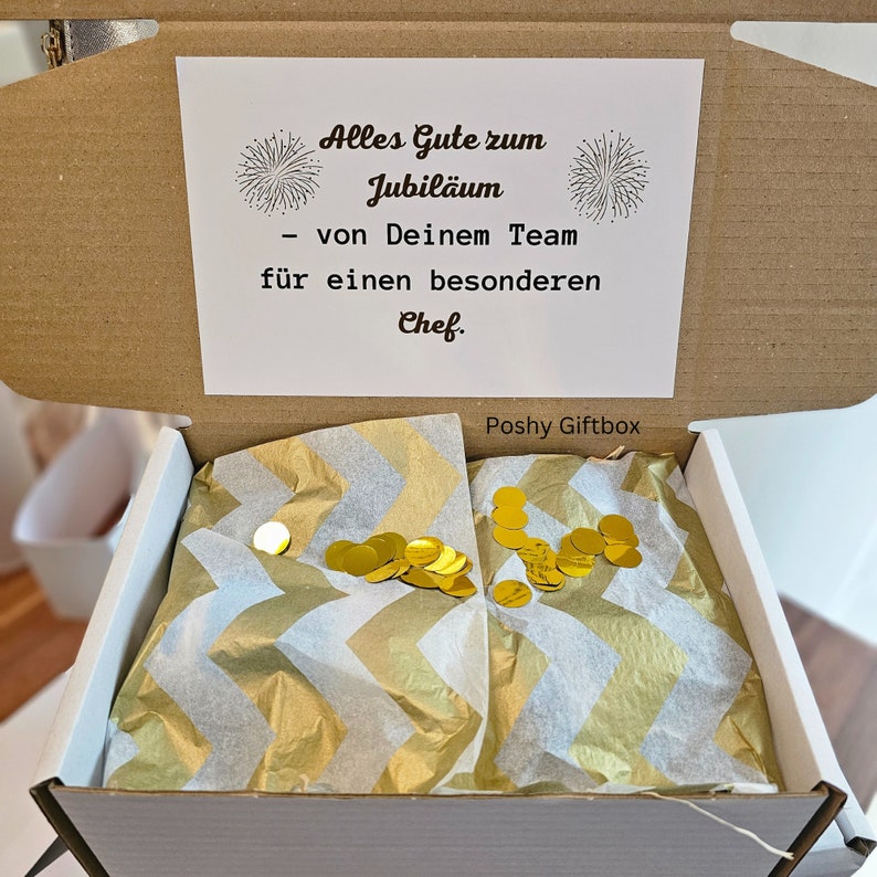 May include: Open cardboard gift box with a white card that reads "Alles Gute zum Jubil&auml;um - von Deinem Team f&uuml;r einen besonderen Chef." The box is filled with gold confetti and white tissue paper.
