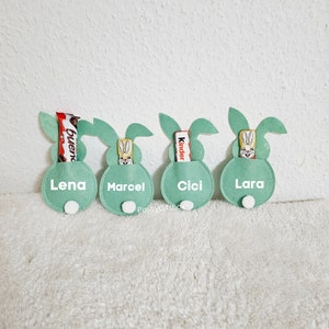 May include: Four green felt bunny-shaped pouches with white names and white bunny faces. Each pouch has a different chocolate bar inside: Bueno, Kinder, and two Kinder.