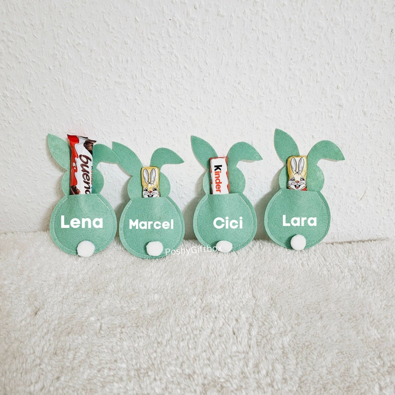 May include: Four green felt bunny-shaped pouches with white names and white bunny faces. Each pouch has a different chocolate bar inside: Bueno, Kinder, and two Kinder.
