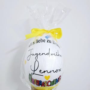 May include: A large white egg-shaped gift wrapped in clear cellophane and tied with a yellow ribbon. The egg has black script that reads "Alles liebe zu Deiner Jugendweihe Lennox &Uuml;berraschung" with yellow heart accents.
