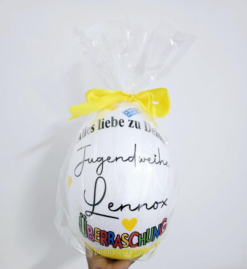 May include: A large white egg-shaped gift wrapped in clear cellophane and tied with a yellow ribbon. The egg has black script that reads "Alles liebe zu Deiner Jugendweihe Lennox &Uuml;berraschung" with yellow heart accents.