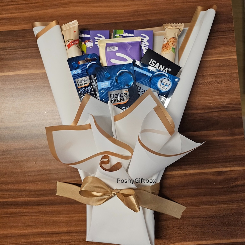 May include: A white gift bouquet with gold ribbon and a variety of items, including chocolate bars, toiletries, and a small package with the text "ISANA" on it. The bouquet is tied with a gold ribbon and has the text "PoshyGiftbox" on the front.