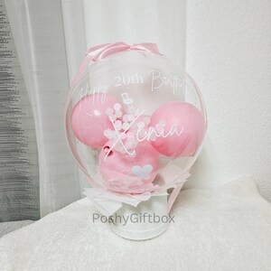 Op de afbeelding: Een doorzichtige bubbelballon met drie roze ballonnen erin, versierd met witte tekst die luidt "Happy 20th Birthday Xenia". De ballon is vastgebonden met een roze lint en staat op een witte container. De woorden "PoshyGiftbox" staan op de container.