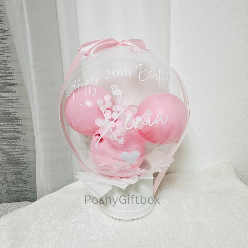 Op de afbeelding: Een doorzichtige bubbelballon met drie roze ballonnen erin, versierd met witte tekst die luidt "Happy 20th Birthday Xenia". De ballon is vastgebonden met een roze lint en staat op een witte container. De woorden "PoshyGiftbox" staan op de container.