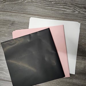 May include: A stack of three square sheets of paper or cardstock arranged in a staggered, overlapping pattern. The top sheet is solid black, the middle sheet is a soft pastel pink, and the bottom sheet is a clean, bright white colour.