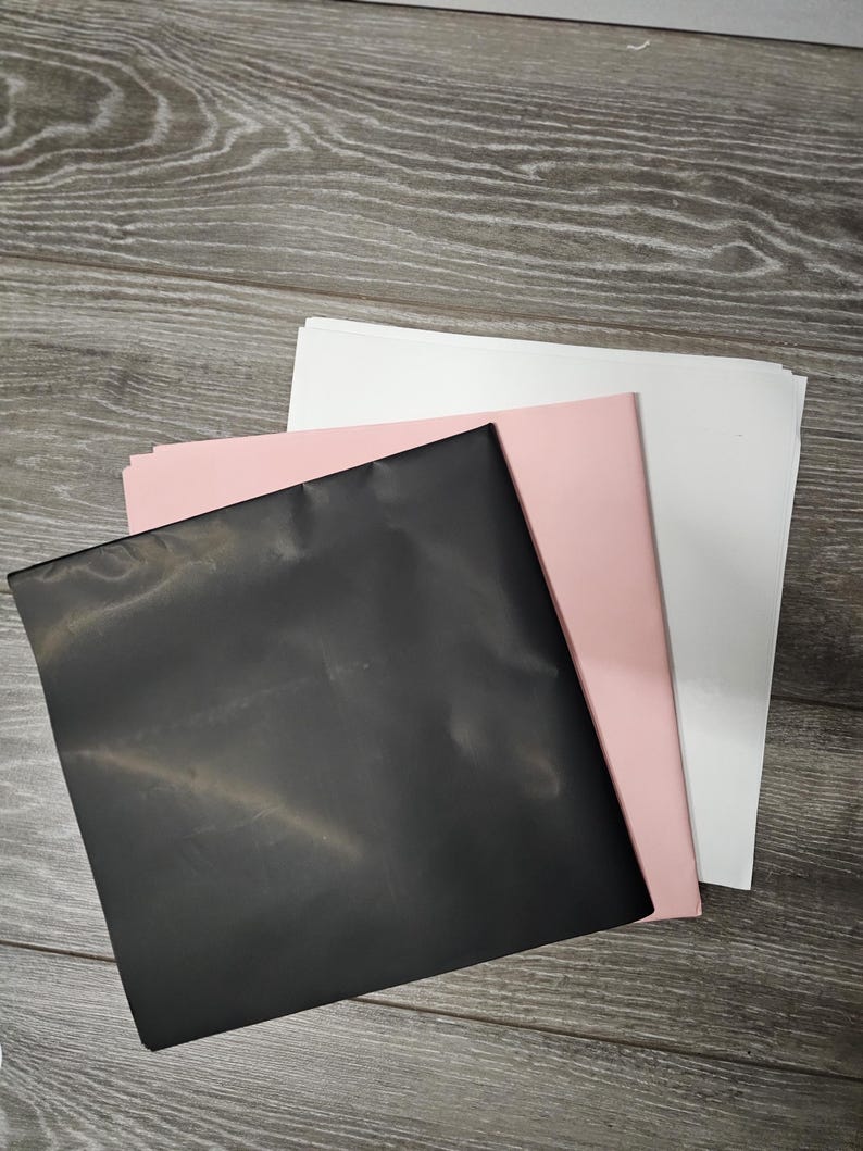 May include: A stack of three square sheets of paper or cardstock arranged in a staggered, overlapping pattern. The top sheet is solid black, the middle sheet is a soft pastel pink, and the bottom sheet is a clean, bright white colour.