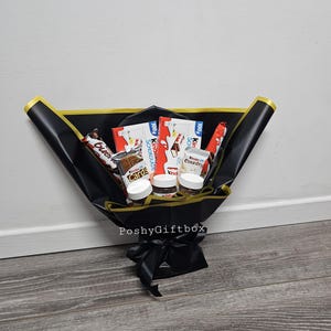 May include: A black gift box with a gold ribbon and a variety of chocolate bars and jars of Nutella inside. The box is tied with a black ribbon and the words "PoshyGiftbox" are printed on the front.