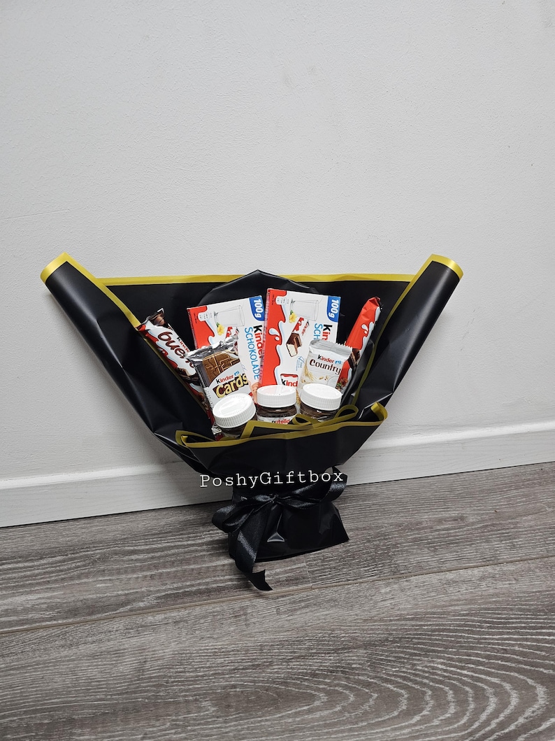 May include: A black gift box with a gold ribbon and a variety of chocolate bars and jars of Nutella inside. The box is tied with a black ribbon and the words "PoshyGiftbox" are printed on the front.