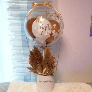 May include: A clear balloon containing two brown balloons and one white balloon with white confetti, featuring gold cursive text that reads Happy Birthday Mama. The balloon is attached to a white cylindrical base filled with dried pampas grass.