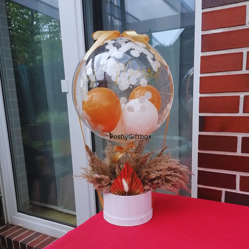 May include: A clear balloon filled with white confetti and smaller orange and white balloons, featuring the gold text "Just Married" and "PoshyGiftbox". The balloon is attached to a white cylindrical box base filled with dried pampas grass.