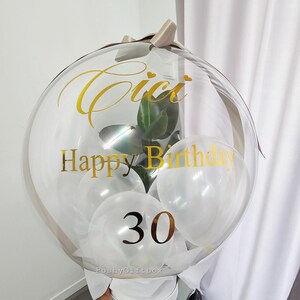 Op de afbeelding: Een doorzichtige bubbelballon met gouden letters met de tekst "Cici Happy Birthday 30". De ballon bevat witte ballonnen en een kleine plant. Hij is vastgebonden met een beige lint en staat op een witte doos.