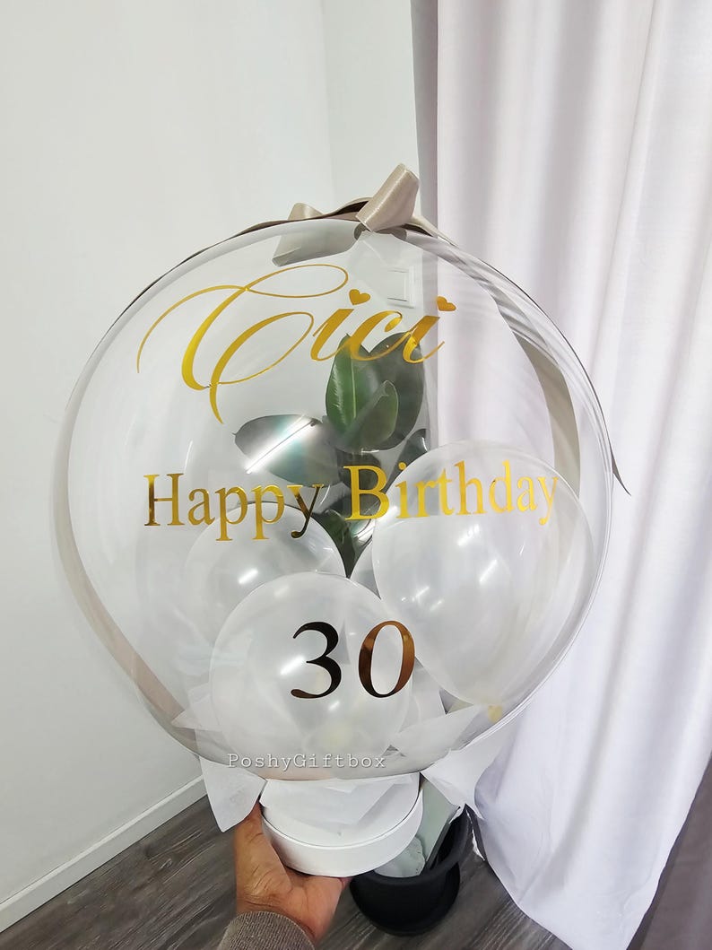 Op de afbeelding: Een doorzichtige bubbelballon met gouden letters met de tekst "Cici Happy Birthday 30". De ballon bevat witte ballonnen en een kleine plant. Hij is vastgebonden met een beige lint en staat op een witte doos.