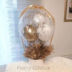 May include: A clear balloon filled with smaller white and gold balloons, featuring the gold text Happy Birthday Michelle 30. The balloon is attached to a white cylindrical base decorated with dried pampas grass and a gold fan-shaped accent.