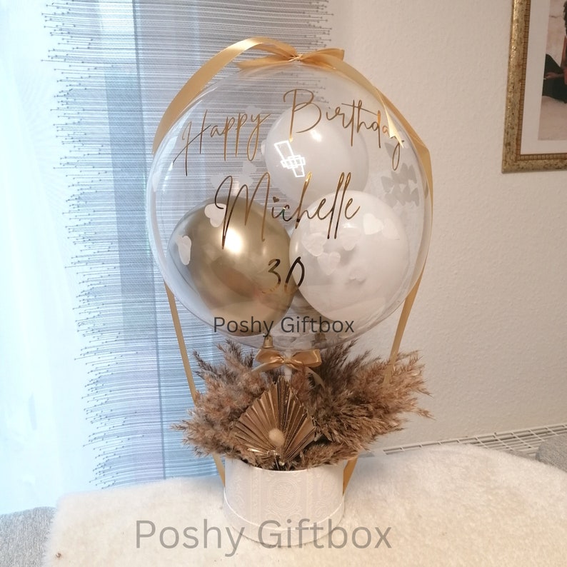 May include: A clear balloon filled with smaller white and gold balloons, featuring the gold text Happy Birthday Michelle 30. The balloon is attached to a white cylindrical base decorated with dried pampas grass and a gold fan-shaped accent.