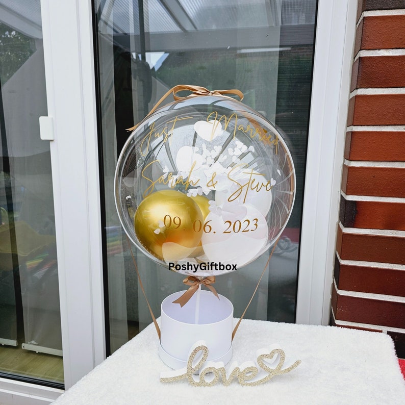 May include: A clear balloon with gold and white balloons inside, tied to a white box with a gold ribbon. The balloon has the text "Just Married Sarah & Steve 09.06.2023 PoshyGiftbox" written on it. A gold glitter "love" sign is in the foreground.