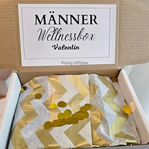 May include: Open gift box with a white and gold chevron patterned tissue paper. The tissue paper is filled with gold confetti circles. The box lid has a white label with the text "M&auml;nner Wellnessbox Valentin Poshy Giftbox".