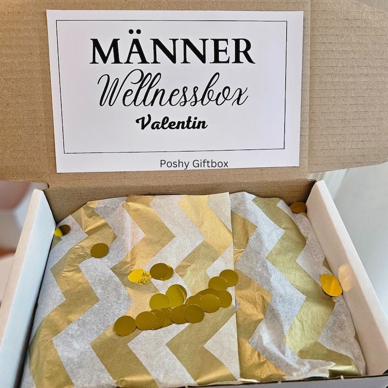 May include: Open gift box with a white and gold chevron patterned tissue paper. The tissue paper is filled with gold confetti circles. The box lid has a white label with the text "M&auml;nner Wellnessbox Valentin Poshy Giftbox".