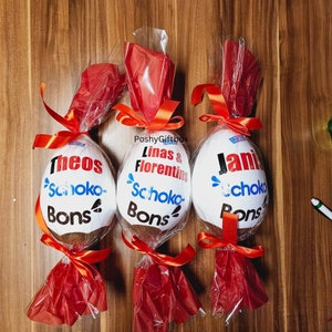 Peut inclure: Trois &oelig;ufs en chocolat blanc envelopp&eacute;s de cellophane rouge avec des n&oelig;uds en ruban orange. Chaque &oelig;uf porte un nom personnalis&eacute; et le texte 'Schoko-Bons' imprim&eacute; dessus.