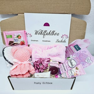 Peut inclure: Une bo&icirc;te-cadeau rose et blanche remplie d'articles de soins personnels, notamment un ours en peluche rose, un bandeau rose, un pouf de bain rose, une bombe de bain rose, une bougie rose, un gommage corporel rose, une lotion corporelle rose, une barre de chocolat rose et une carte rose qui dit "Wohlf&uuml;hlbox Einatmen. Ausatmen. L&auml;cheln."